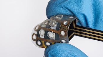 Manos con guantes azules de nitrilo sujetan una lámina flexible e inclinada con circuitos dorados y pequeños sensores circulares.