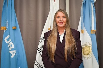 Una mujer con traje oscuro y cabello largo posa frente a la bandera azul de la Universidad de Buenos Aires y la bandera de Argentina