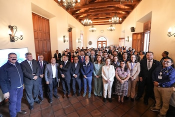 Personas reunidas en un salón con una mesa directiva, un orador en un podio, un intérprete de lengua de señas, banderas y una pantalla