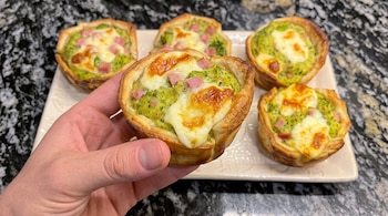 Una mano sostiene una tartaleta de masa rellena de brócoli, queso y jamón frente a una bandeja con cuatro piezas más.