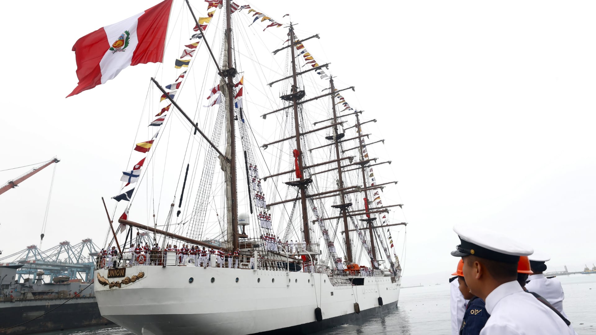 El buque escuela B.A.P. Unión de la Marina de Guerra del Perú navega con su tripulación en cubierta, banderas de diversas naciones y la bandera peruana.