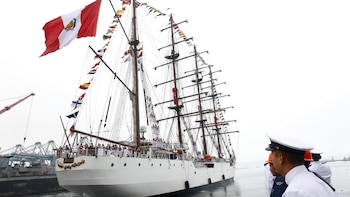 Gran buque velero blanco con bandera peruana y banderas de señales, tripulación en cubierta. Oficiales de uniforme blanco en primer plano y grúas de puerto