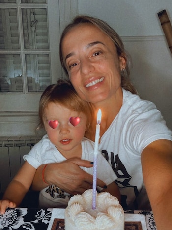 Una mujer sonriente abraza a una niña frente a un pastel blanco con una vela encendida, con los ojos de la niña cubiertos por corazones rojos