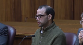 Un hombre con anteojos y suéter verde permanece sentado en un tribunal frente a un micrófono