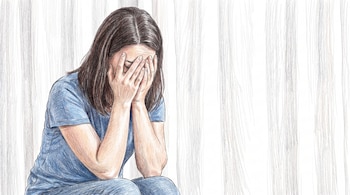 Una persona sentada con el rostro cubierto por las manos. Viste camiseta y pantalones azules. Fondo claro con líneas verticales.
