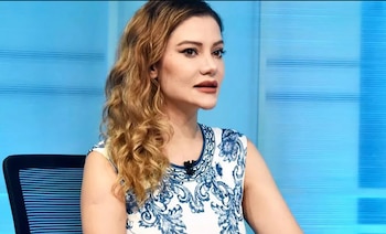 Mujer con cabello ondulado, top blanco con estampado azul y maquillaje sentada en una silla negra con respaldo azul frente a un fondo celeste