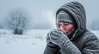 Persona con abrigo y gorro cubiertos de nieve, sosteniendo una taza de la que sale vapor, con mejillas enrojecidas, en un paisaje invernal nevado.