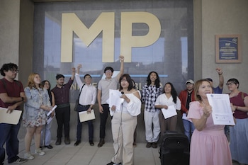 Grupo de personas frente a un edificio gris con un logo dorado "MP". Varias personas tienen los puños en alto, dos mujeres sujetan micrófonos y documentos