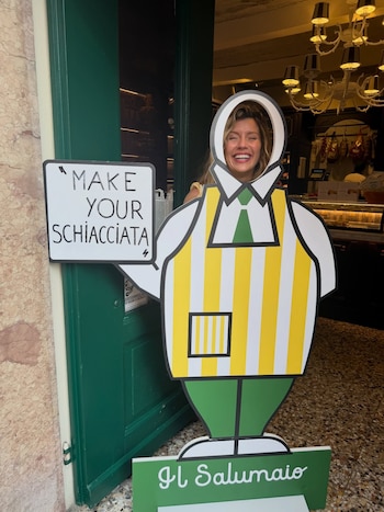 Mujer sonriendo dentro de un cartel de cartón con uniforme a rayas amarillas y blancas. Se ven letreros con “Make your schiacciata” e “Il Salumaio”
