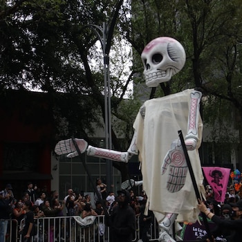 Una figura gigante de esqueleto vestida de blanco avanza sostenida por varas frente a una multitud detrás de vallas metálicas