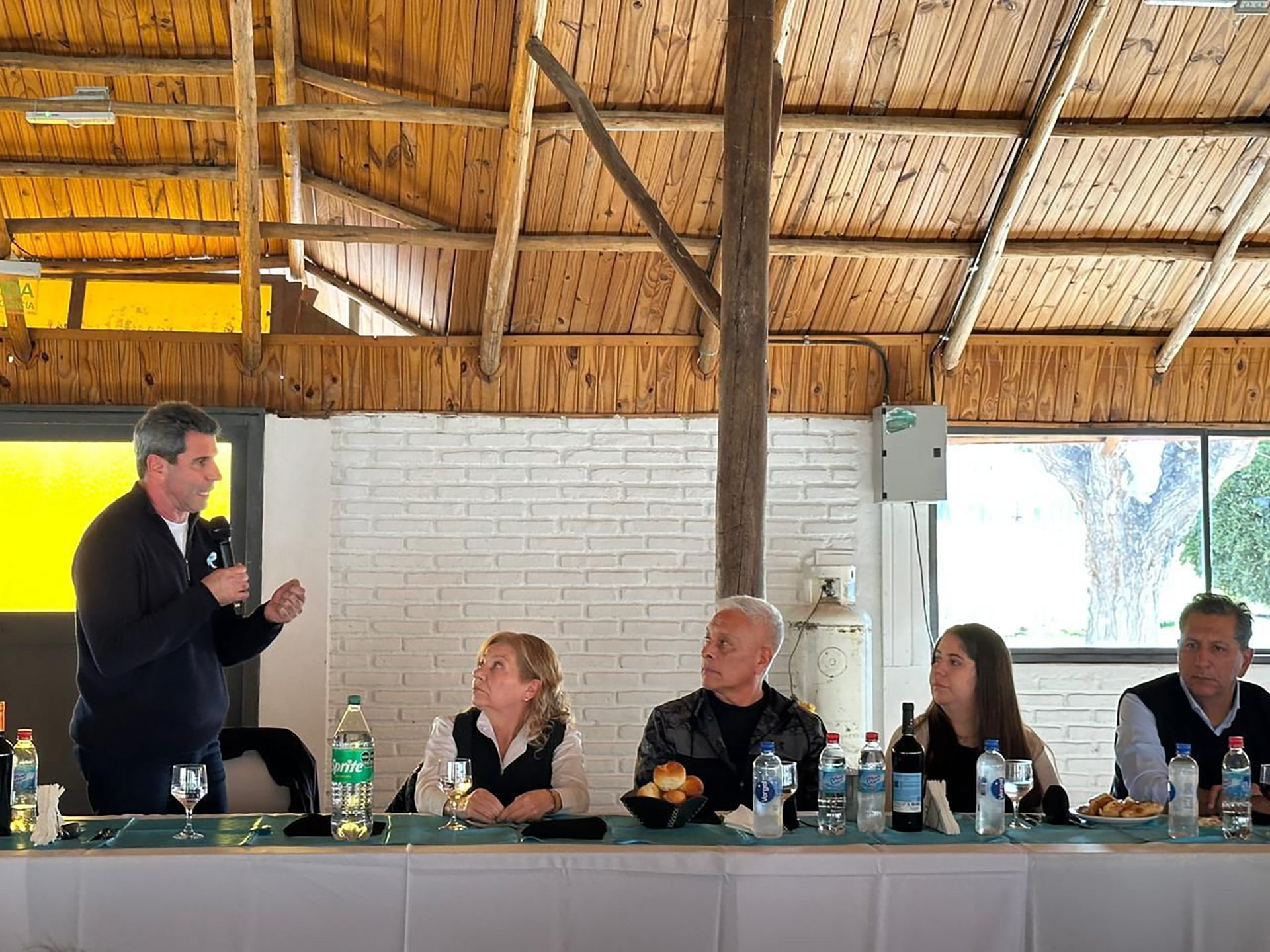 Sergio Uñac, con un micrófono, se dirige a cuatro personas sentadas en una mesa con bebidas y panecillos, en un salón con techo de madera y paredes de ladrillo blanco.