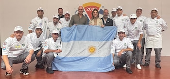 Un grupo de diecisiete personas, la mayoría hombres y dos mujeres, en uniformes de chef, posan sonrientes con la bandera argentina en un fondo blanco