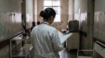 Vista posterior de una mujer con bata blanca y estetoscopio que lee un expediente en un pasillo de hospital.