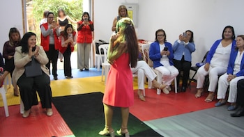 Joven con vestido rosa de espaldas sobre piso multicolor, frente a un grupo de mujeres sentadas. Algunas aplauden y otras graban con teléfonos