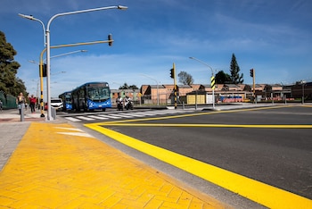 Una acera amarilla precede a una intersección asfaltada con paso de peatones, semáforos, autobuses azules y motocicletas en circulación