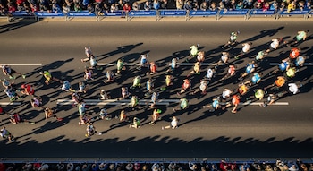 Vista aérea de múltiples corredores de maratón en una calle asfaltada, con largas sombras proyectadas por el sol y espectadores tras vallas.