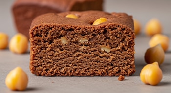 Un trozo de brownie de garbanzo cortado muestra su textura interna densa con pequeñas inclusiones. Varios garbanzos crudos se dispersan sobre una superficie clara.