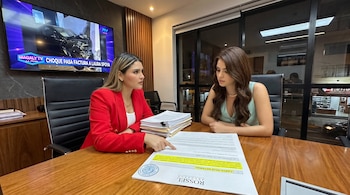Dos mujeres sentadas en una mesa de madera revisan un documento legal en una oficina con un televisor encendido y una pila de papeles.