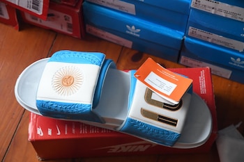 Primer plano de sandalias blancas y celestes con el Sol de Mayo y el número 10 dorado, sobre una caja Nike roja, con cajas Adidas al fondo