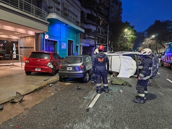 Vista nocturna de un accidente vial. Tres vehículos, uno volcado, en una calle urbana. Dos rescatistas y una ambulancia en el lugar. Escombros en asfalto