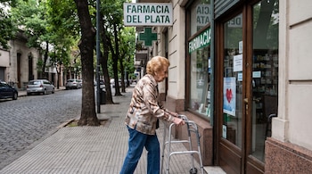Una mujer con blusa estampada y pantalones vaqueros camina con un andador de metal frente a la entrada de una farmacia.