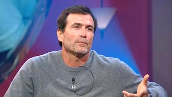 Un hombre con suéter gris gesticula con la mano en un estudio con iluminación azul y violeta