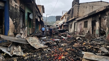 Una calle comercial en Sonsonate, El Salvador, con edificios calcinados y escombros humeantes tras un incendio devastador, mientras personas caminan entre los restos.