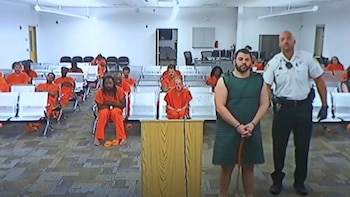 Hombre con barba y uniforme verde junto a un agente uniformado en sala de tribunal. Detrás, varias personas en monos naranjas se sientan en bancos