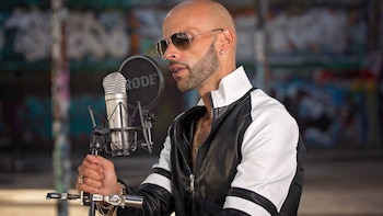 Un hombre con barba, gafas de sol y chaqueta deportiva sostiene el soporte de un micrófono de condensador con filtro antipop
