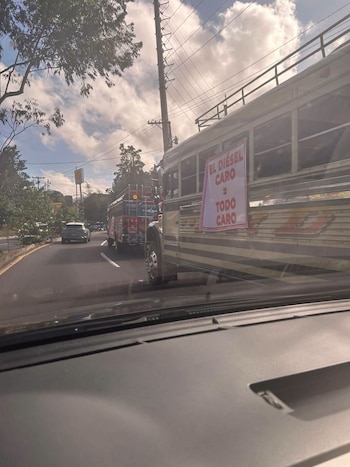 Vista desde el interior de un vehículo hacia un autobús en la vía con un cartel que dice EL DIÉSEL CARO = TODO CARO