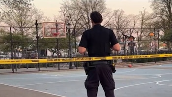 Un oficial de policía de espaldas con uniforme oscuro en una cancha de baloncesto; cinta amarilla de "escena del crimen" atraviesa el plano medio