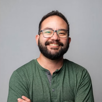 Hombre sonriente con barba, gafas y camiseta verde sobre un fondo gris neutro