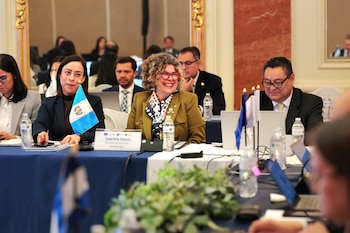 Grupo de personas sentadas en una mesa de conferencia con banderas de Guatemala, laptops, botellas de agua, carteles de nombres y una planta