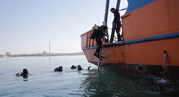 Varios buzos con trajes de neopreno y equipo suben por una escalera a un barco naranja. Otros buzos flotan en el agua. Fondo con costa y una chimenea