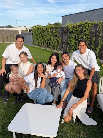 Grupo familiar grande en un jardín con césped verde. Se ven adultos, jóvenes y dos bebés sonrientes, con una mesa blanca en primer plano y plantas trepadoras al fondo