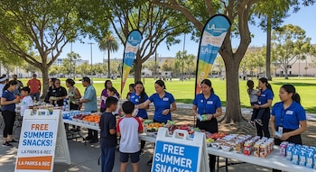 Niños y adultos reciben refrigerios como frutas, jugos y snacks de personal en mesas al aire libre en un parque verde con árboles y carteles del programa.