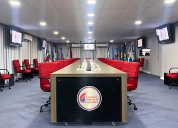 Mesa de conferencias con sillas rojas, micrófonos, el logotipo de la Comisión Federal de Impuestos, pantallas y banderas de fondo
