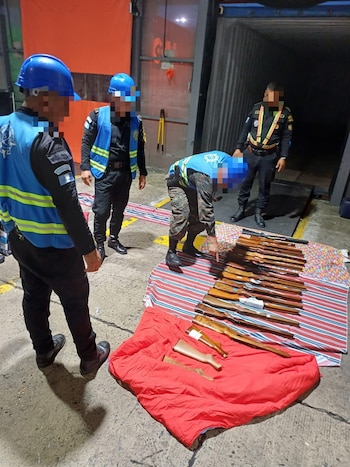 Interior de un almacén con varias personas en cascos azules y chalecos reflectantes. Inspeccionan y organizan cajas de cartón apiladas en palets