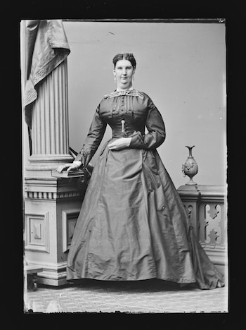 Retrato en blanco y negro de mujer con vestido largo y aretes. Posa de pie junto a una columna con tela y un libro. Una balaustrada con urna decora el fondo