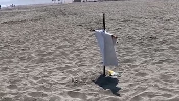 Una playa de arena clara con un poste vertical, una tela blanca colgando y una botella de plástico en la base. Personas y estructuras al fondo