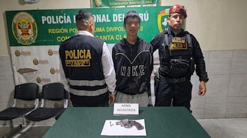 Un hombre con los ojos cubiertos permanece entre dos policías frente a una mesa con un revólver y un cartel de evidencia.