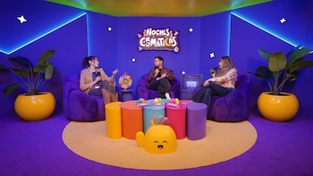 Tres personas sentadas en sillones morados con micrófonos en la mano, en una escenografía con mesas de colores y plantas