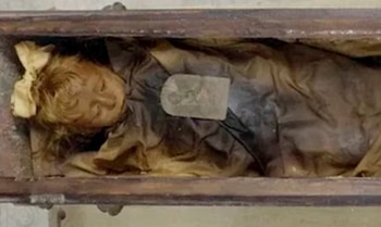 Cuerpo momificado de una niña dentro de un ataúd de madera. Tiene cabello claro, un lazo en la cabeza, viste ropa oscura y está envuelta en telas marrones. Una placa rectangular descansa sobre su pecho