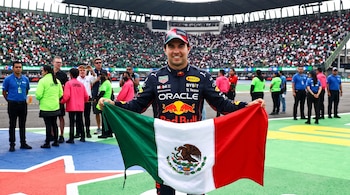 Sergio Pérez con el uniforme de Red Bull sostiene una bandera de México desplegada en una pista de carreras con público al fondo.