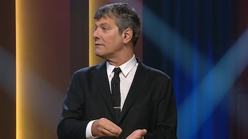 Hombre de cabello gris, traje oscuro, camisa blanca y corbata negra, gesticula con las manos en un escenario iluminado con tonos amarillos y azules