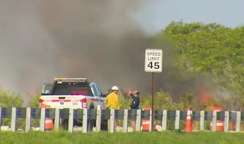 Camioneta de bomberos y personal cerca de un incendio forestal. Humo denso y llamas visibles. Un cartel de límite de velocidad de 45 y conos de tráfico en primer plano