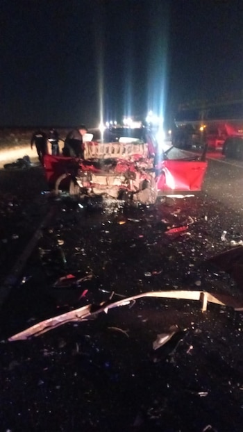 Fotografía nocturna de un vehículo rojo gravemente dañado en una carretera oscura, rodeado de escombros. Luces en el fondo y siluetas de personas son visibles
