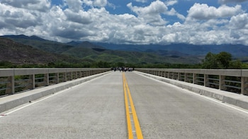 Vista frontal de un largo puente de concreto con doble línea amarilla, un grupo de personas en el extremo, montañas y cielo nublado de fondo