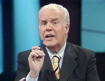 Primer plano del periodista Julio Ricardo, un hombre de cabello blanco, vestido con traje oscuro y corbata dorada y azul, sosteniendo un bolígrafo azul