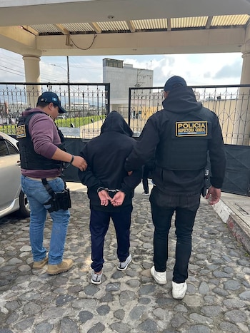 Dos policías con chalecos antibalas escoltan a una persona con sudadera negra y esposada, caminando sobre un empedrado frente a un portón metálico